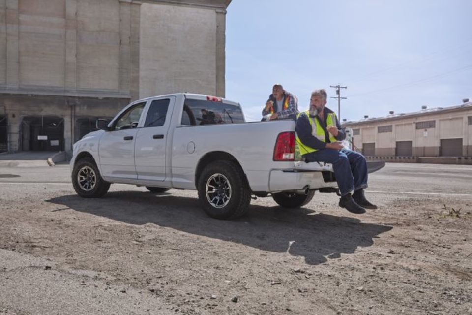 two men in construction vests sit in the bed of a white truck eating lunch at their construction work site